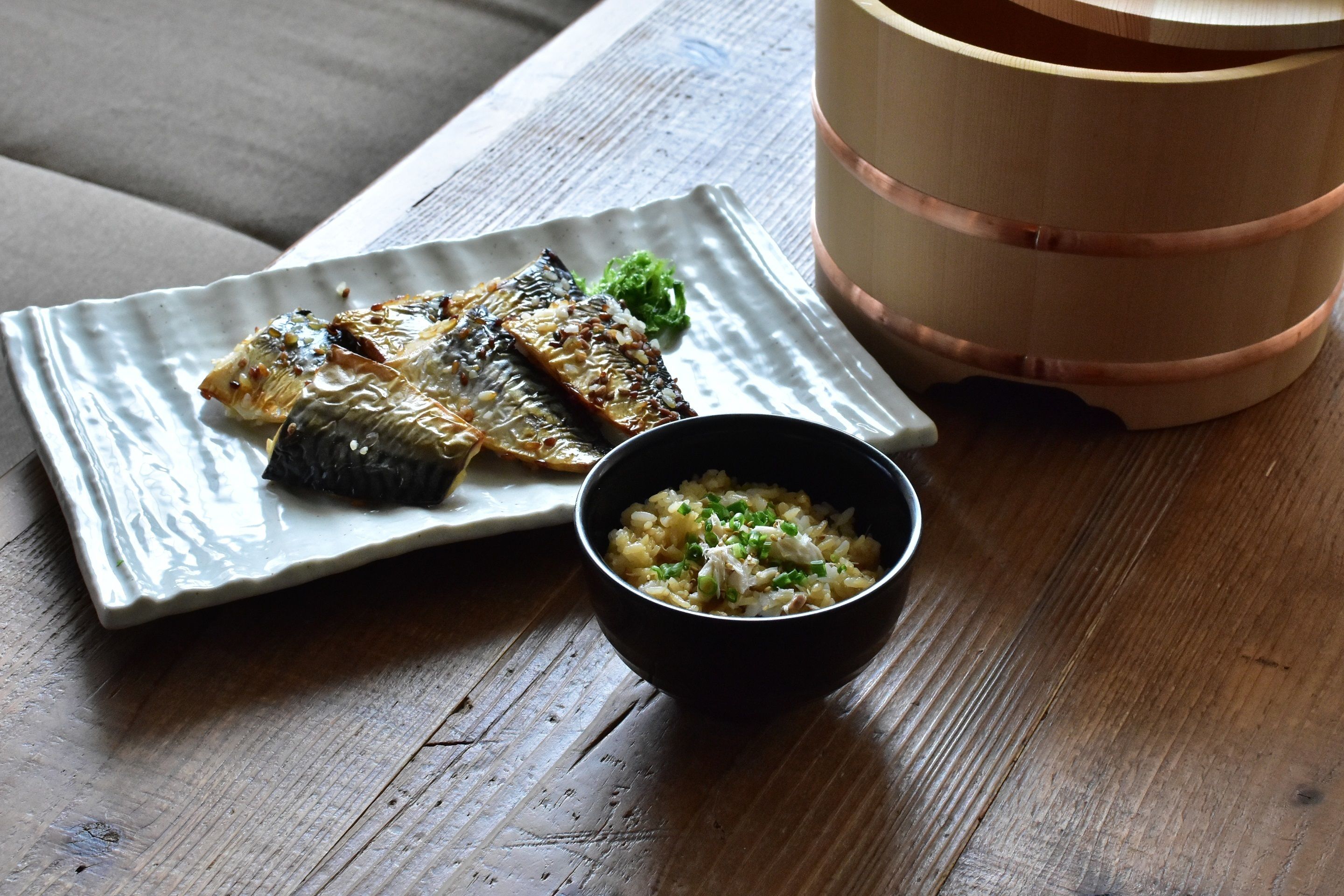 A plate of grilled fish and a bowl of rice with green onions on a wooden table.