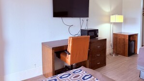 Une chambre d’hôtel dotée d’un téléviseur à écran plat fixé au mur, d’un bureau en bois avec une chaise, d’une petite armoire en bois, d’une lampe et d’un lit recouvert d’une couvre-lit à motifs.