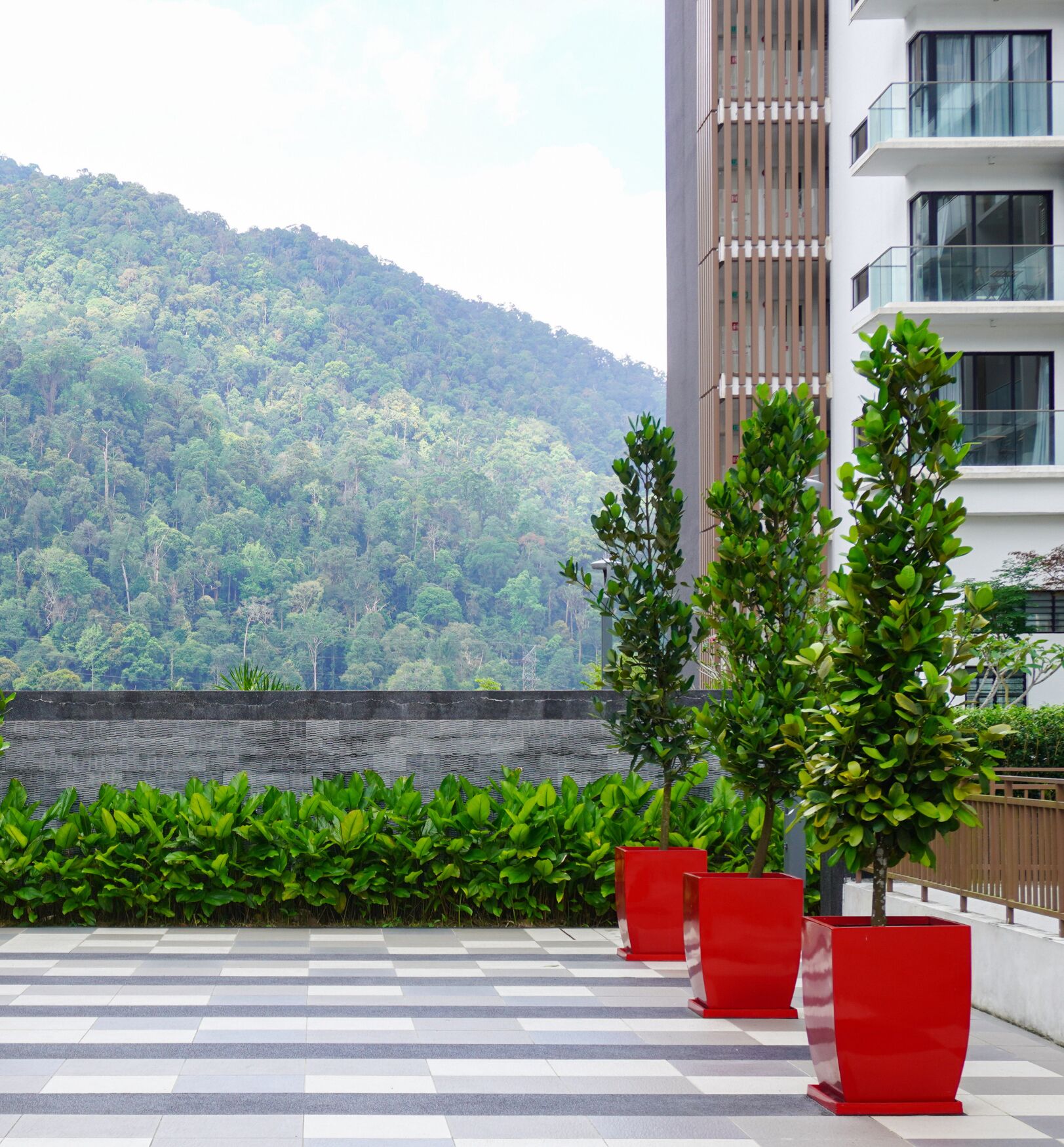 A rooftop terrace with a view of a forested hill, red planters, and a modern building with balconies.