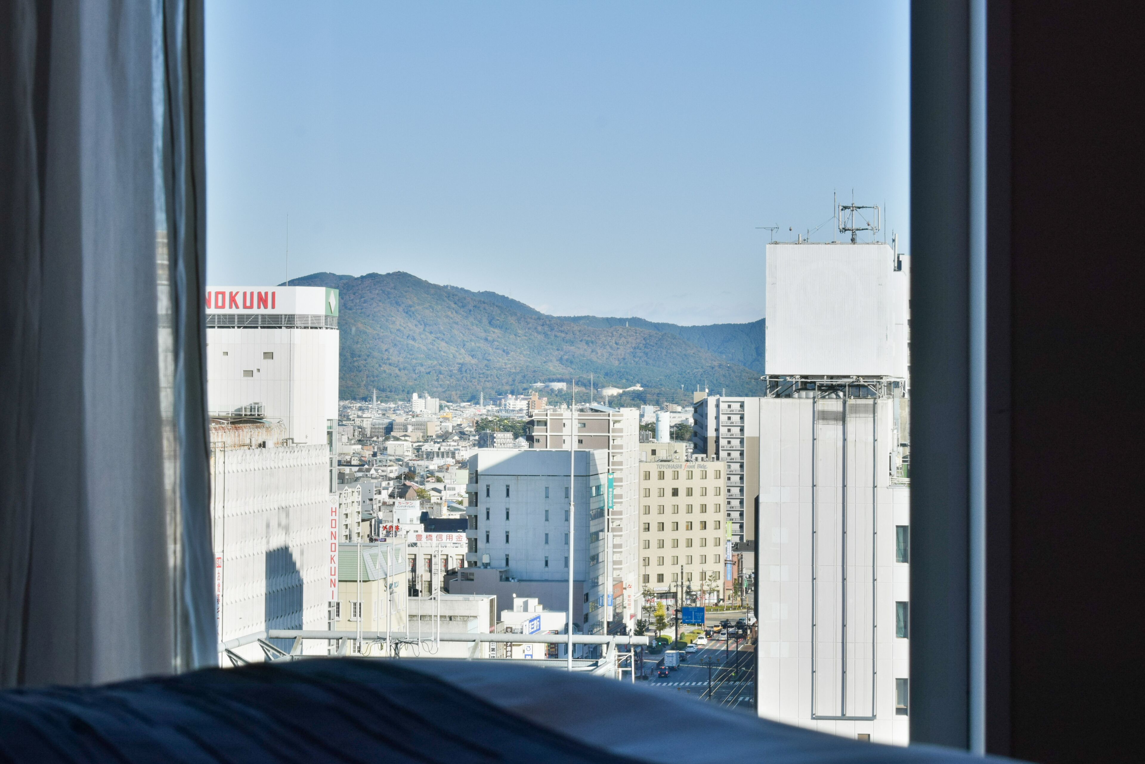 A cityscape with a view of mountains through a window, featuring buildings with signage including "NOKUNI".