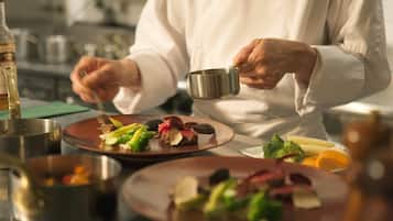 A chef in a white uniform holding a small metal bowl, with plates of food in the foreground.
