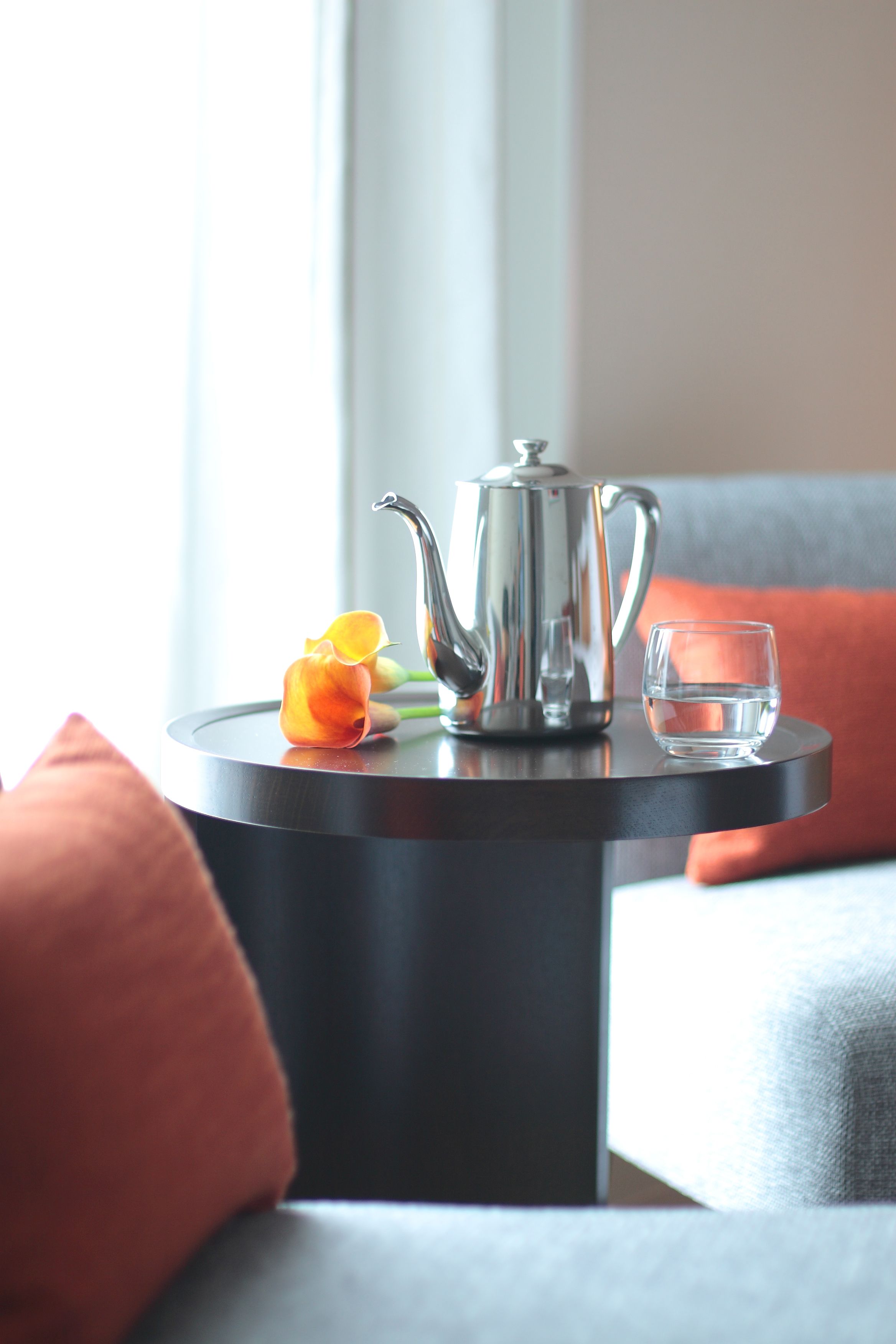 A small round table with a silver teapot, a glass of water, and a single orange flower.