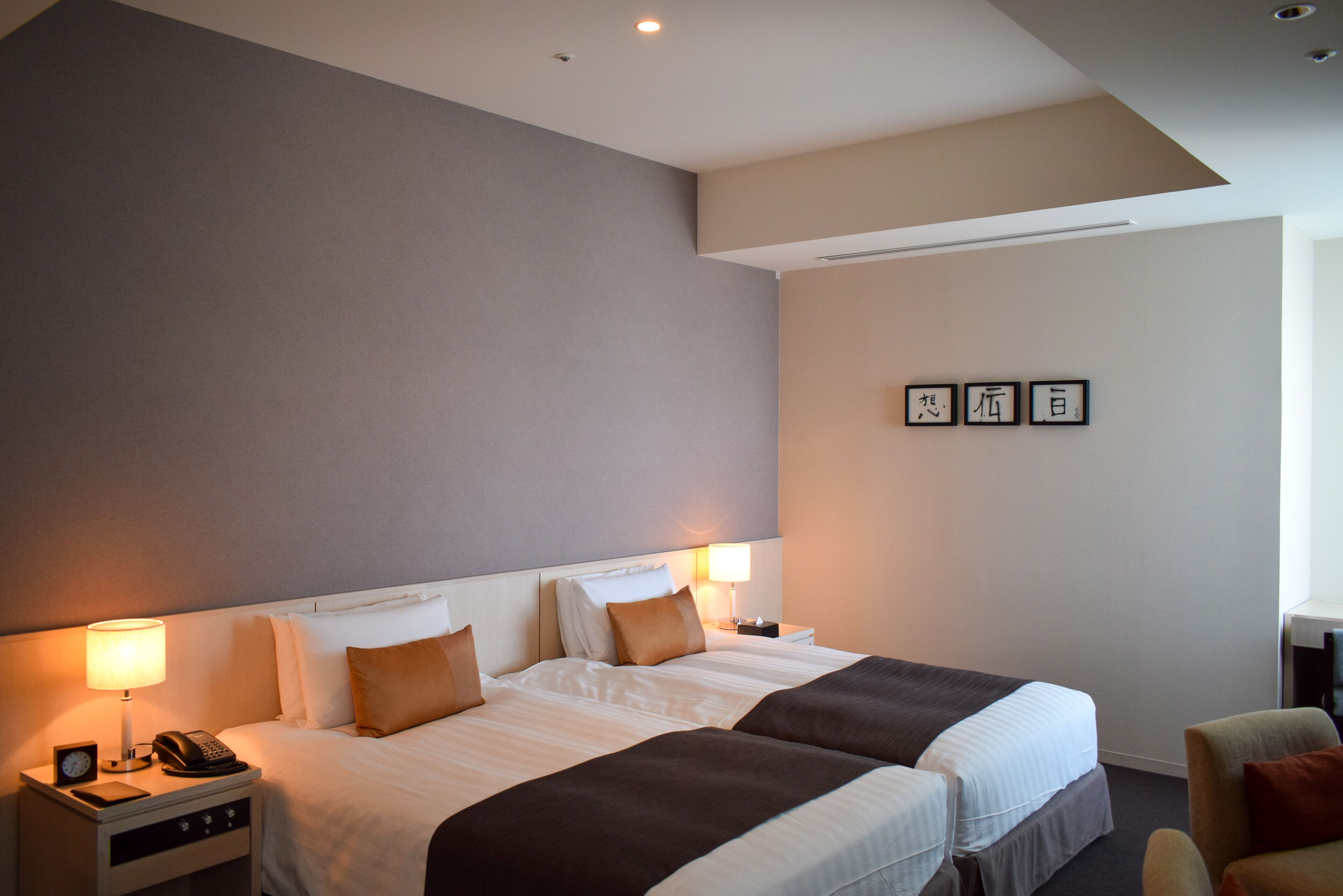 A hotel room with two beds, a nightstand with a lamp and phone, and a wall with framed calligraphy.