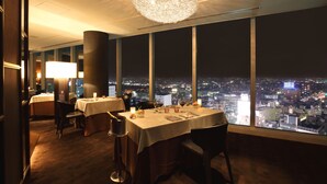 Un restaurante con mesas dispuestas para comer, una araña y vistas de la ciudad a través de amplios ventanales.