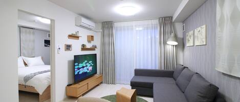 A modern living room with a sofa, a TV, a dining table, and a bed in the background.