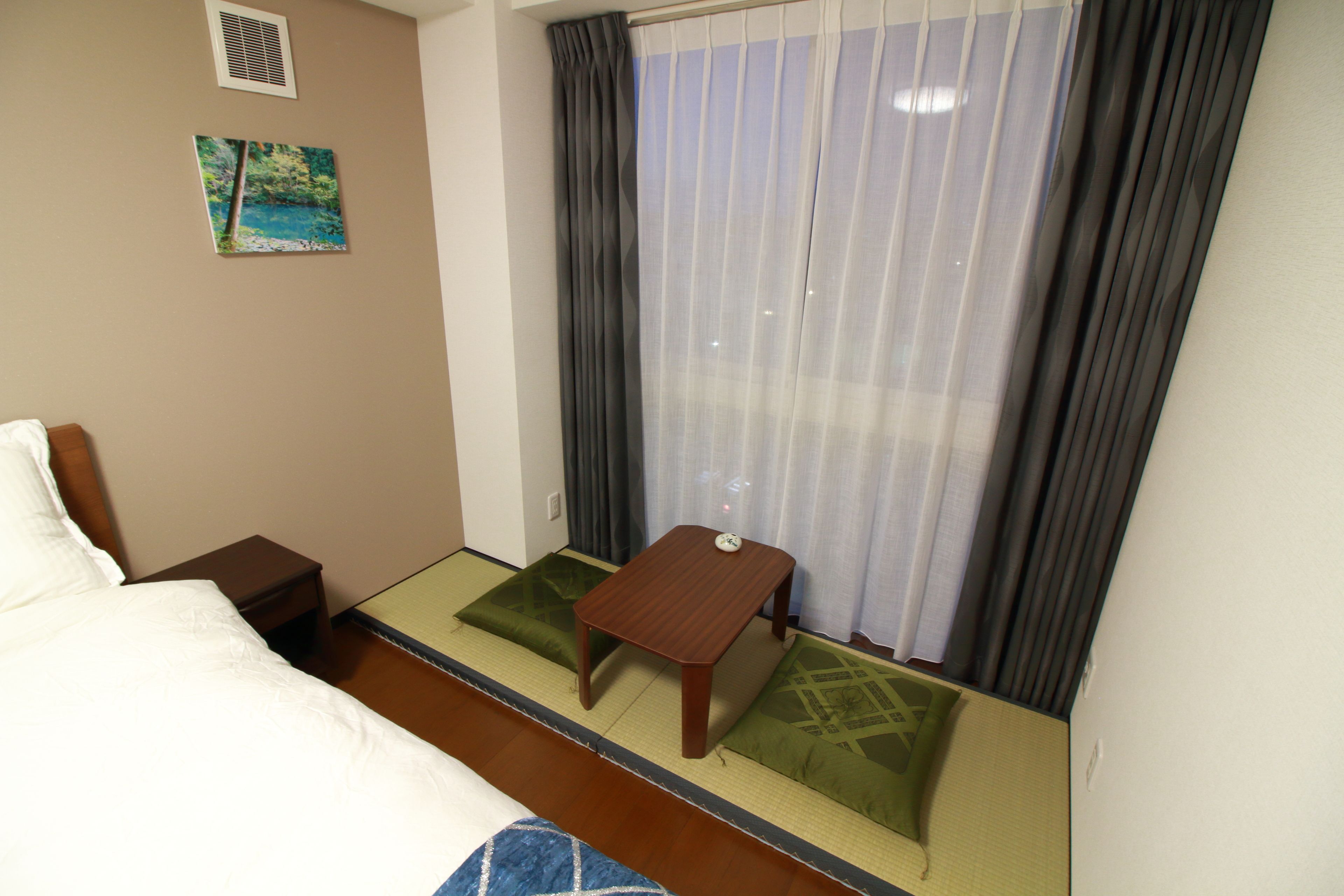 Una camera d'albergo con un letto, un tavolino in legno, un futon con cuscini verdi, un quadro appeso alla parete e una finestra con tende.