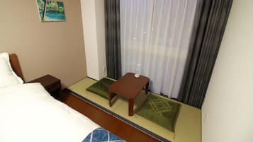 Una camera d'albergo con un letto, un tavolino in legno, un futon con cuscini verdi, un quadro appeso alla parete e una finestra con tende.