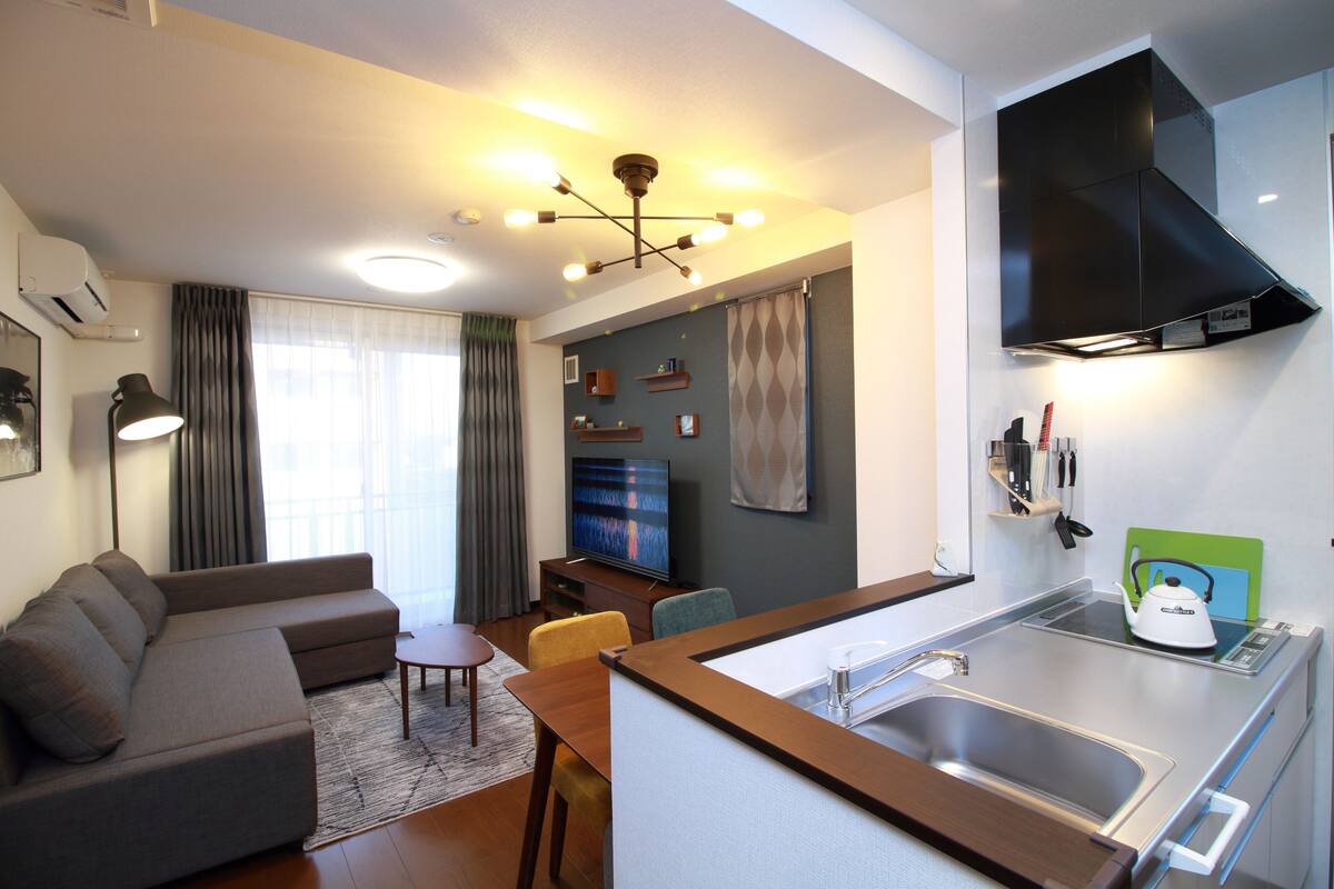 A modern living room with a sofa, a dining table, and a kitchen area with a sink and kettle.