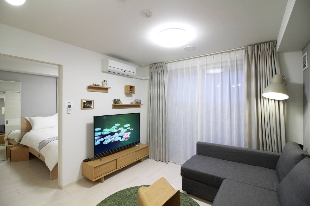 A modern living room with a flat-screen TV, a grey sofa, a wooden TV stand, a small wooden table, and a green rug.