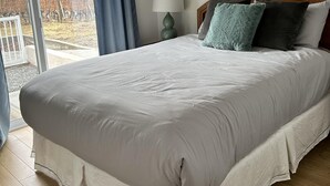 A bedroom with a large bed, a wooden headboard, a bedside table with a lamp, and a view of a vineyard through a sliding door. - Sonora Desert Inn (Osoyoos)
