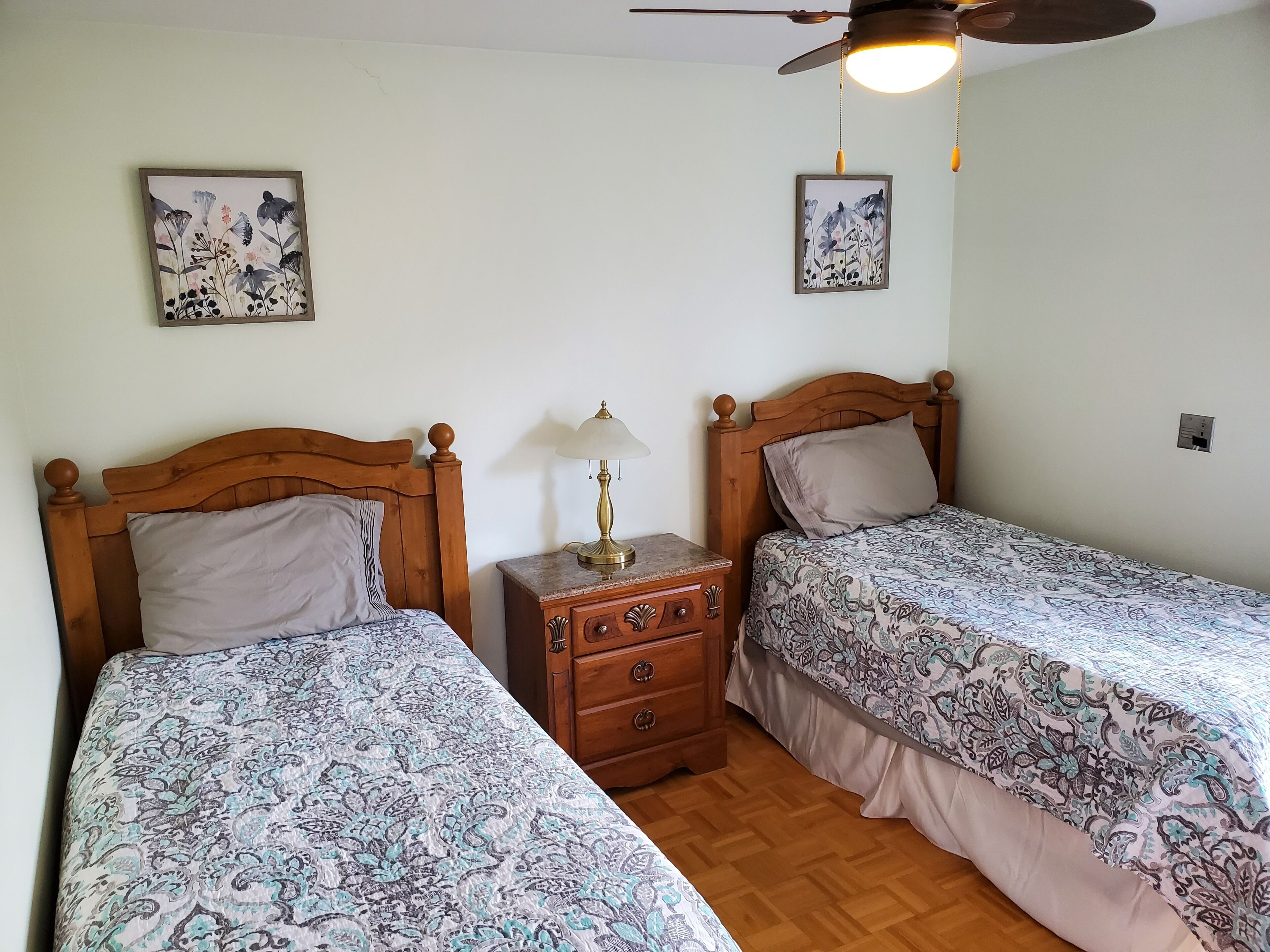 Una habitación con dos camas, cabeceros de madera, una mesita de noche con lámpara y decoración floral en la pared.
