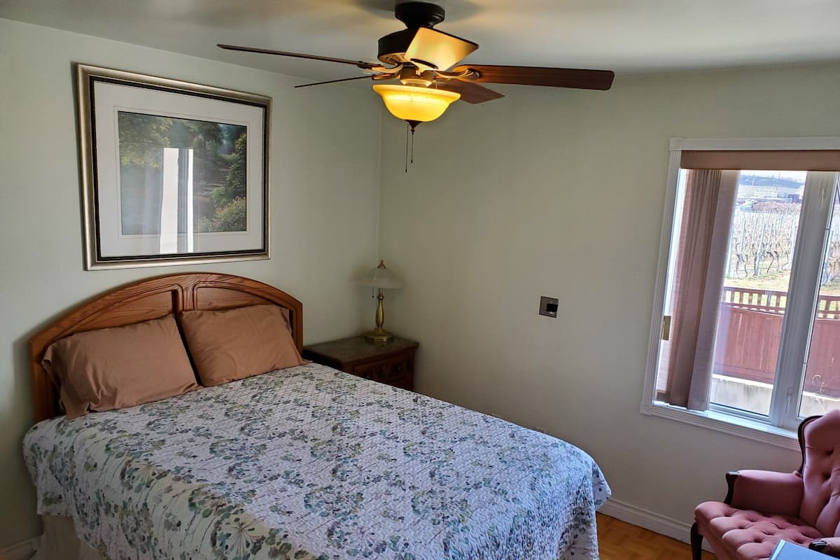 Une chambre à coucher comprenant un lit, un ventilateur de plafond, une table de chevet, une lampe, un tableau encadré, une fenêtre et une chaise.