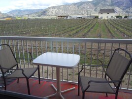Un balcón con dos sillas y una mesa, con vistas a un viñedo con hileras de vides y un edificio al fondo.
