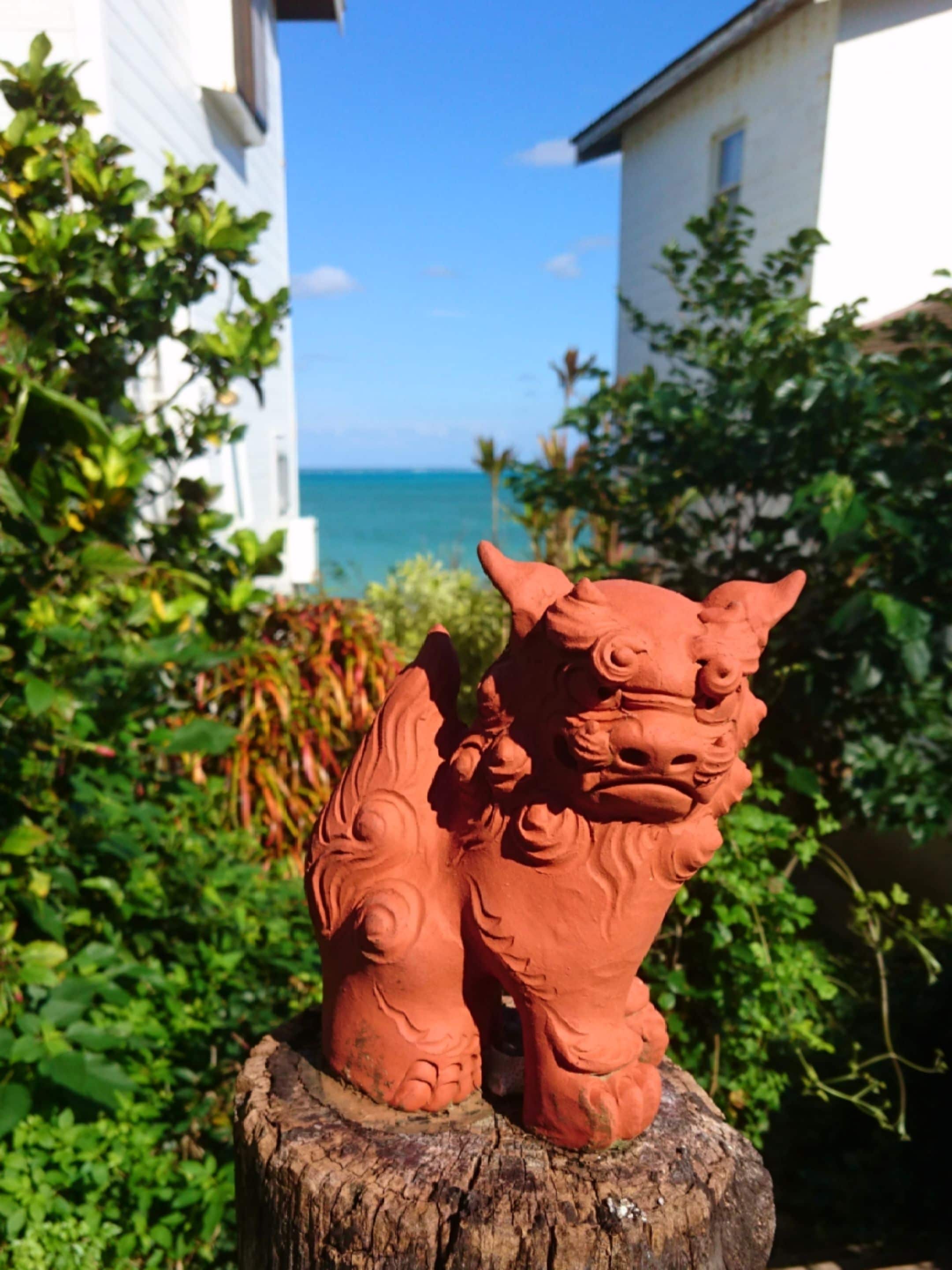 Eine leuchtend rote Löwenstatue auf einem Baumstumpf, im Hintergrund ein weißes Gebäude und der Ozean.