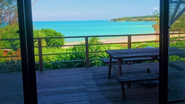 A wooden deck with a table and bench, overlooking a beach with clear turquoise waters and a distant shoreline. - Private Beach House (Motobu)