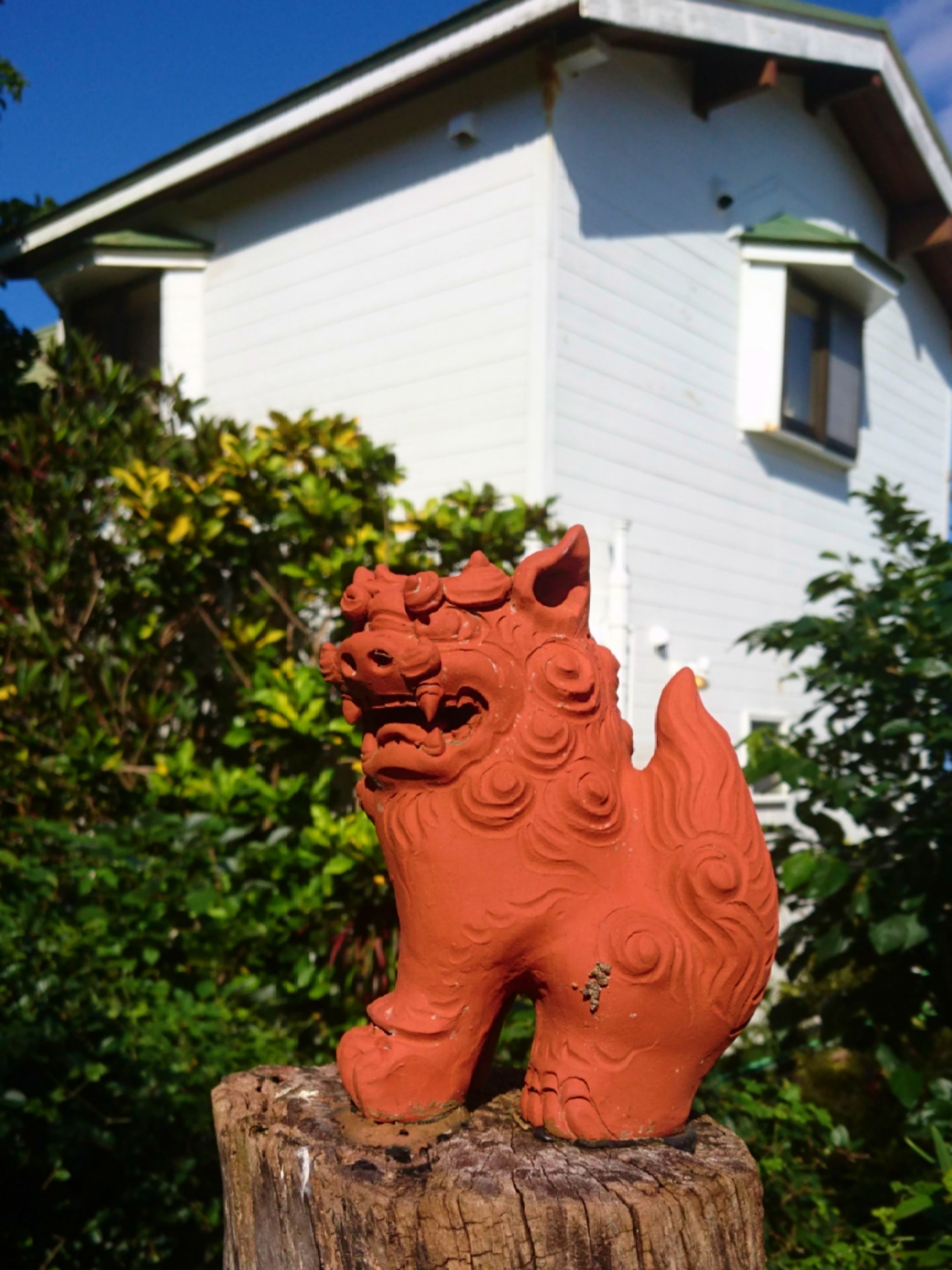 A terracotta lion statue on a wooden post, with a white building and green foliage in the background.