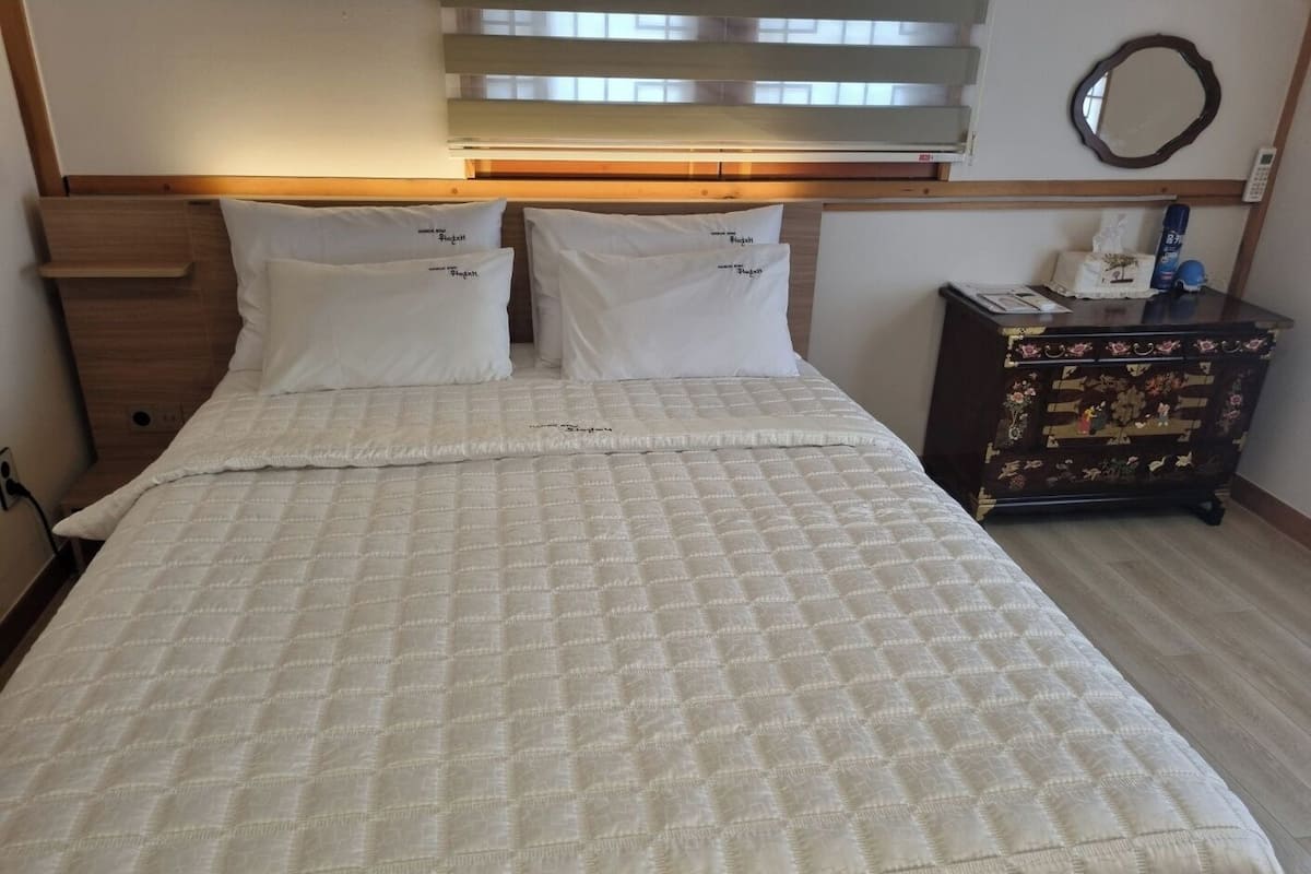 A neatly made bed with a quilted white duvet, a wooden headboard, and a bedside table with a floral-patterned dresser.