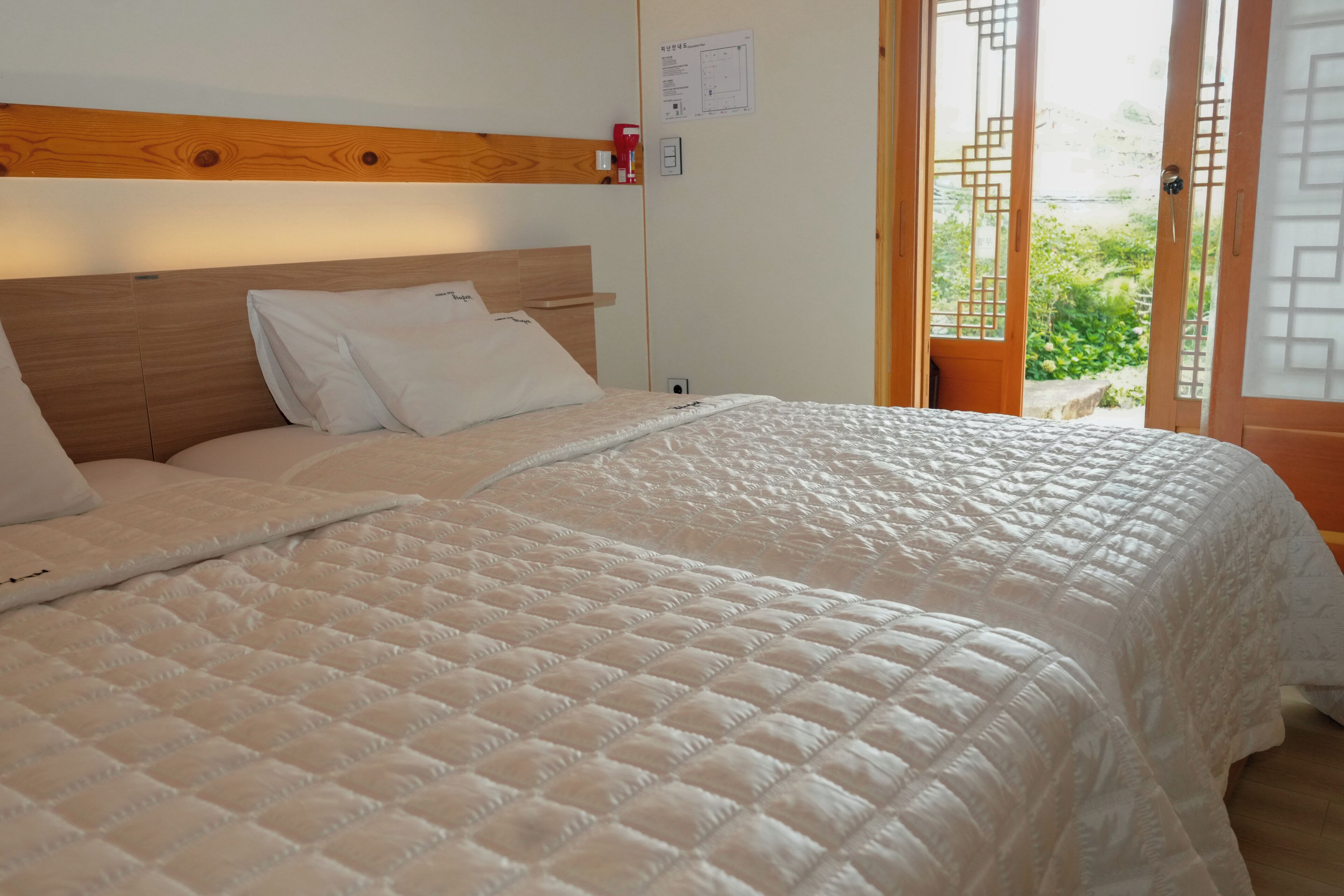 A neatly made bed with a quilted white duvet, a wooden headboard, and a view of the outdoors through a door with a geometric design.