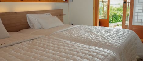 A neatly made bed with a quilted white duvet, a wooden headboard, and a view of the outdoors through a door with a geometric design.