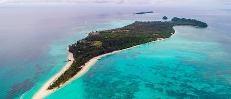 An aerial view of a tropical island with a narrow beach, clear turquoise waters, and lush greenery.