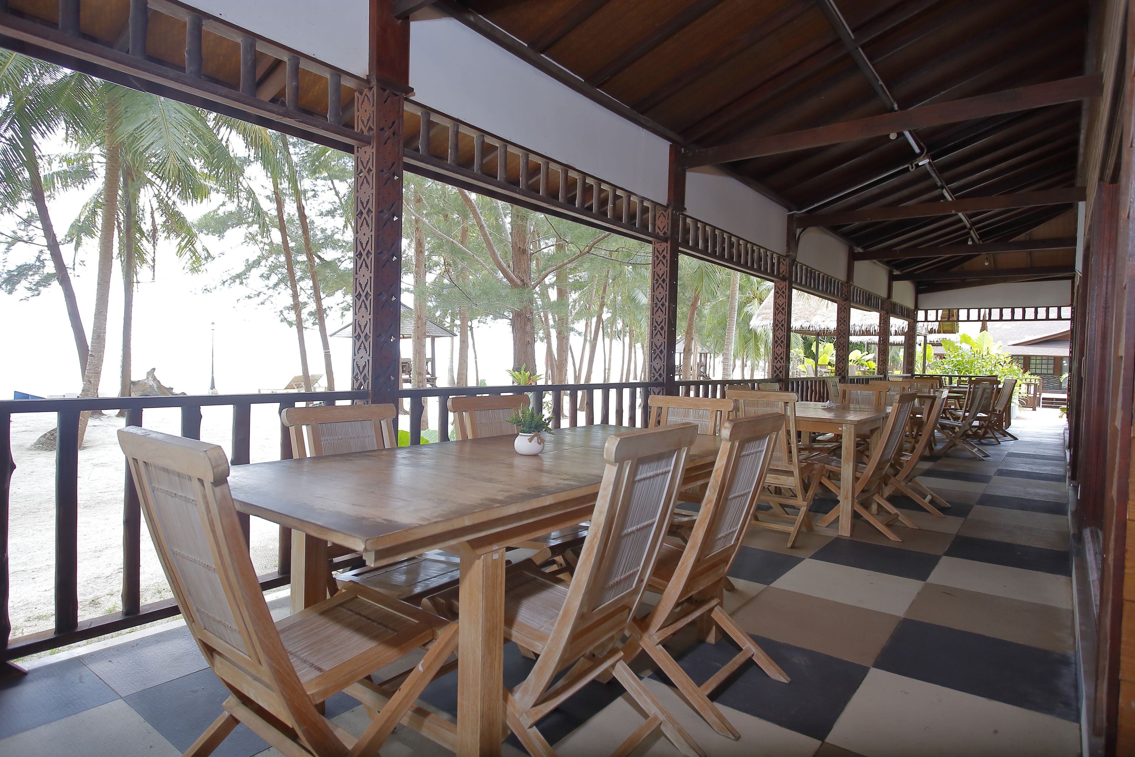 A covered outdoor dining area with wooden tables and chairs, large windows, and a view of palm trees.