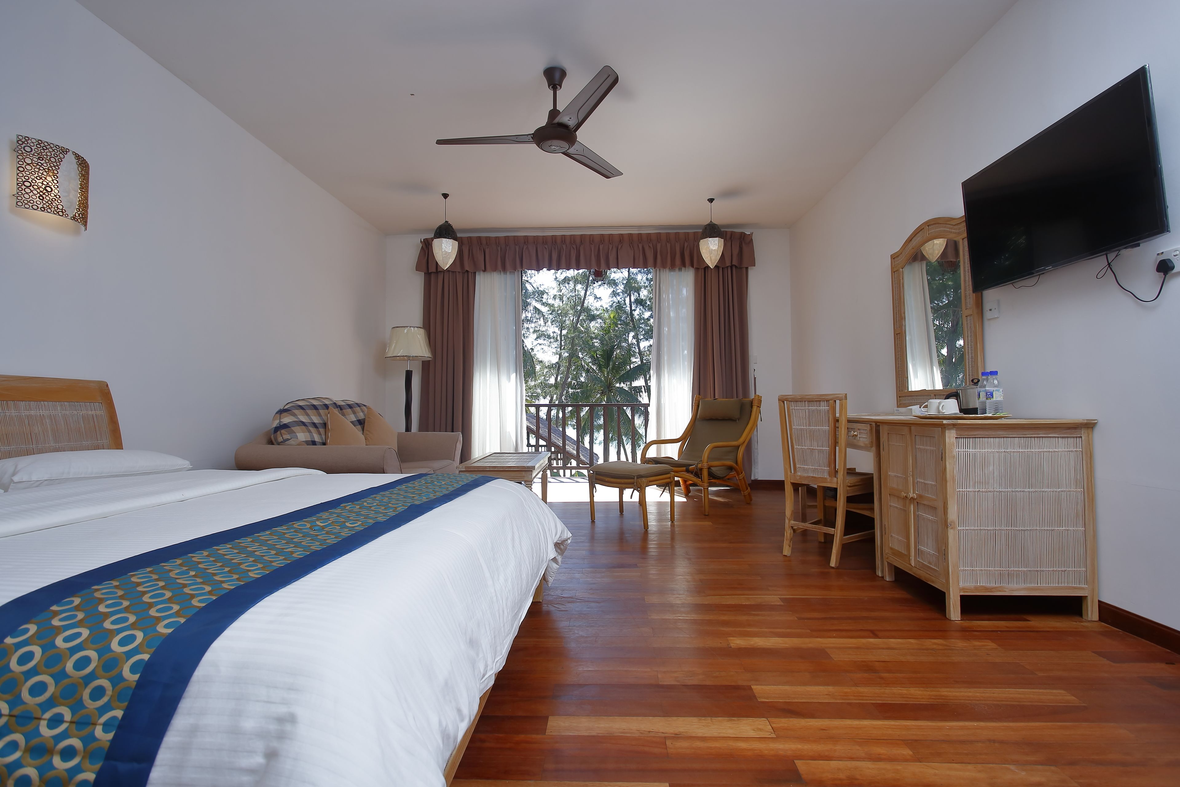A hotel room with a bed, a desk, a chair, a television, and a balcony with a view of trees.
