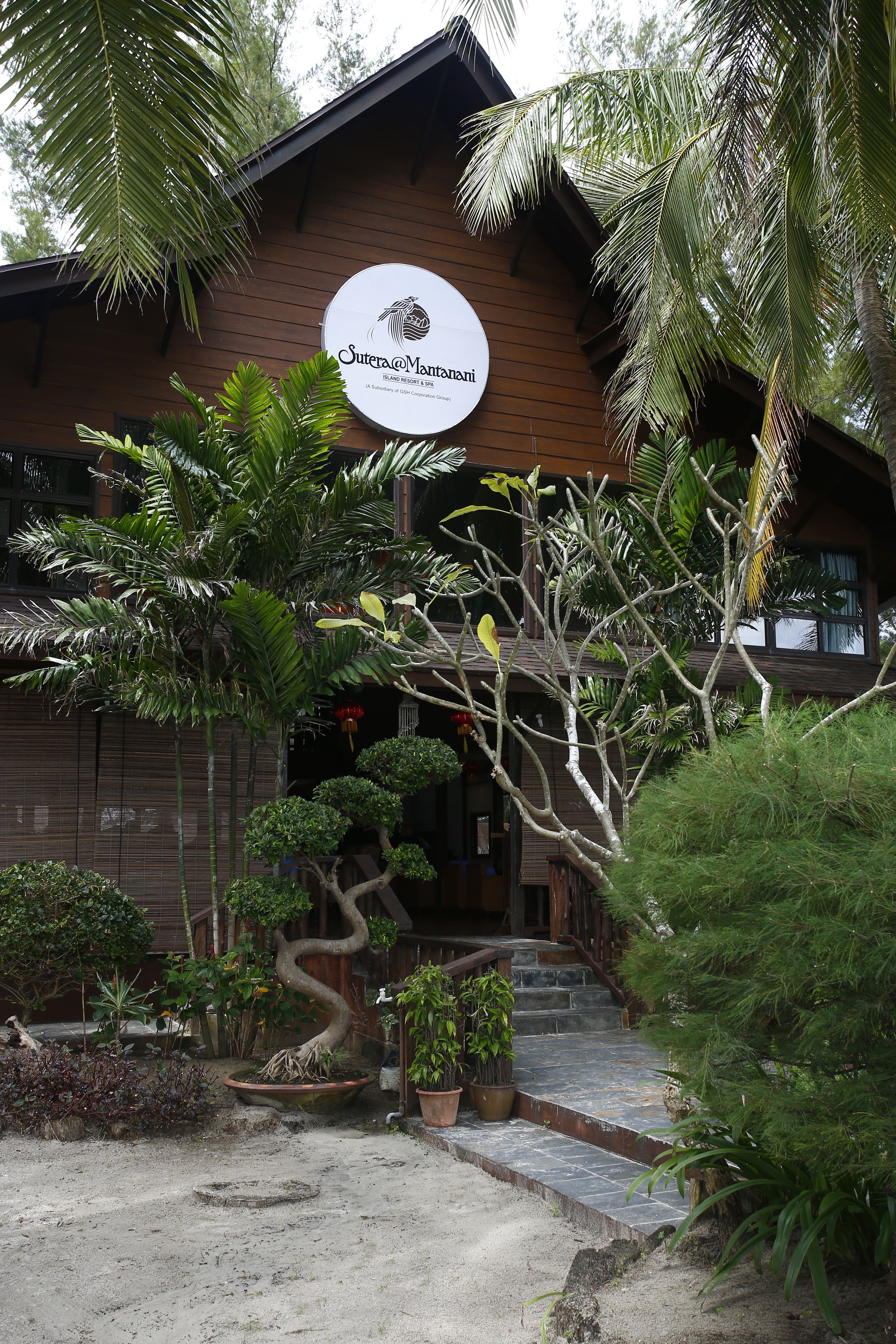 A building with a sign reading "Sutra@Manani" and a well-maintained garden with various plants and a stone pathway.