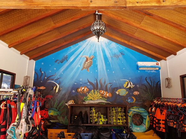 Un espacio interior con techo de madera, un mural de una escena submarina, equipo de buceo y una repisa con varios objetos.