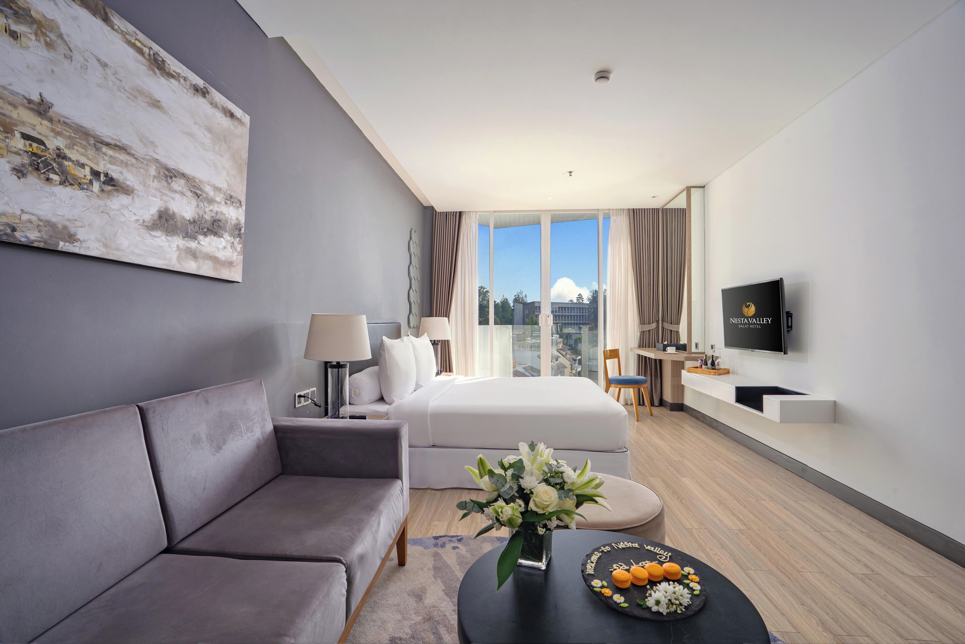 Una habitación de hotel moderna con sofá gris, cama, televisor de pantalla plana y un ventanal con vistas a la ciudad.