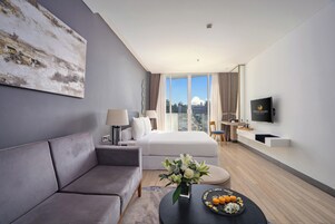 A modern hotel room with a grey sofa, a bed, a flat-screen TV, and a large window offering a city view.