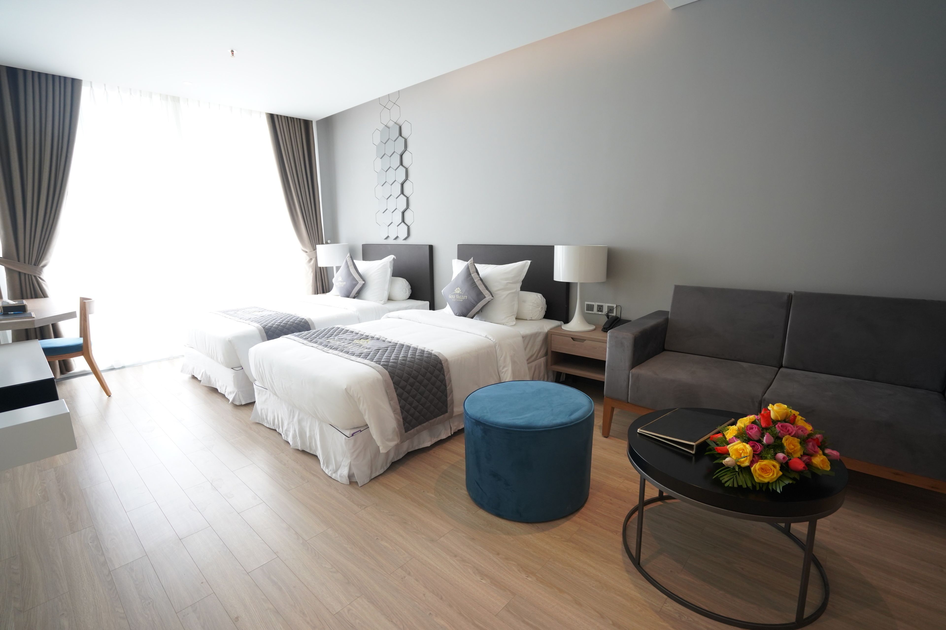 A modern hotel room with a bed, two bedside tables, a sofa, a round ottoman, and a coffee table with flowers.