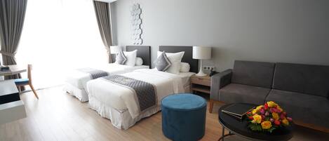 A modern hotel room with a bed, two bedside tables, a sofa, a round ottoman, and a coffee table with flowers.