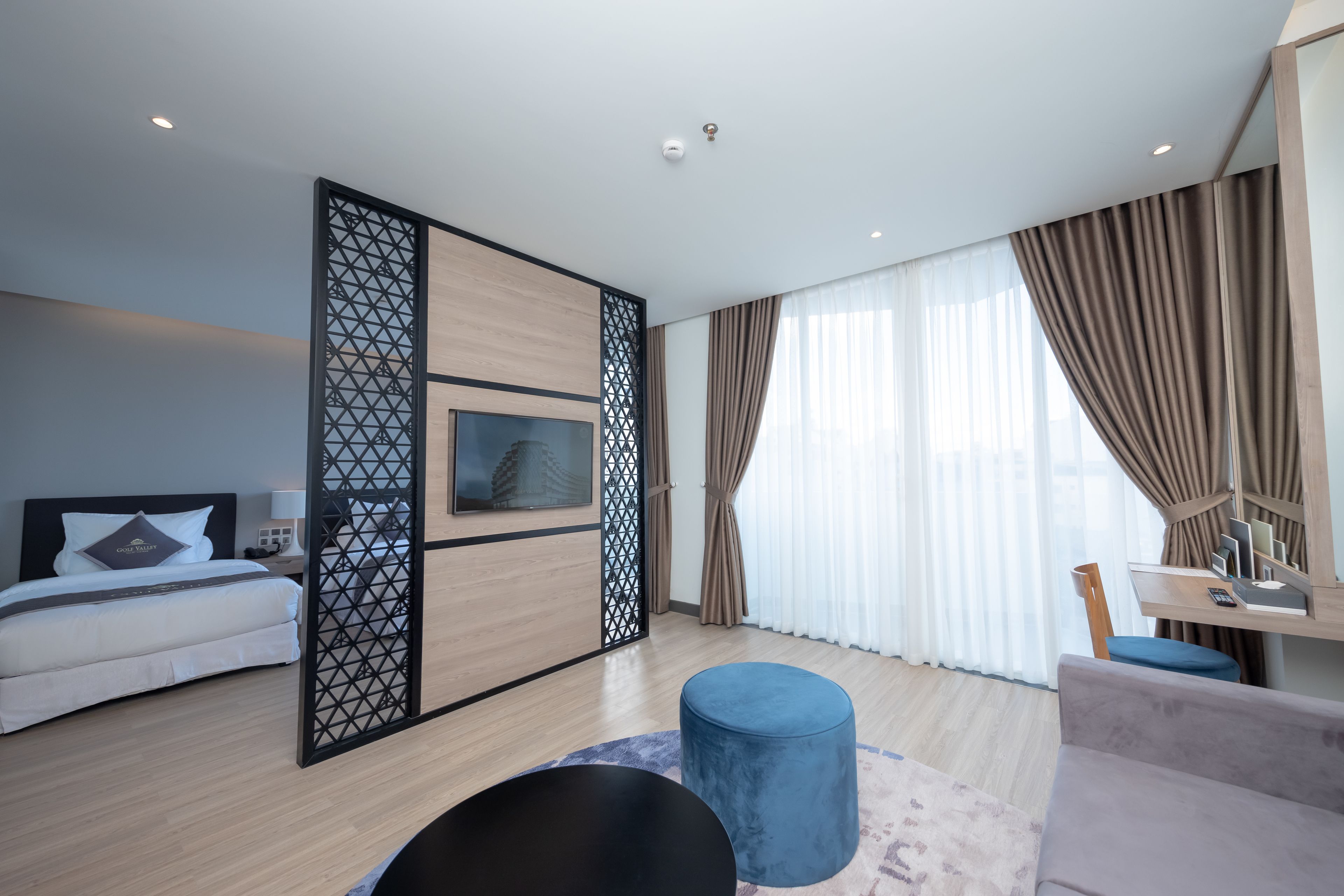 A modern hotel room with a bed, a TV, a desk, and a large window with curtains.