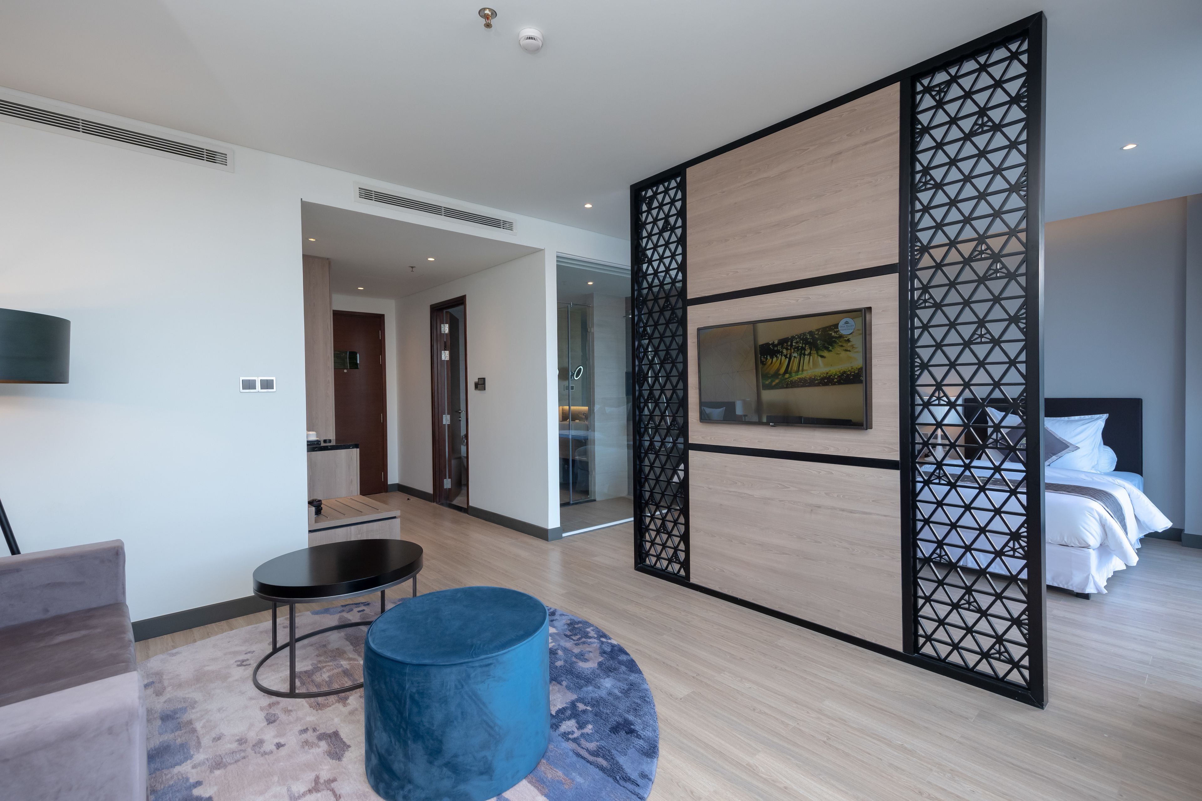 A modern hotel room with a wooden floor, a blue ottoman, a flat-screen TV, a bed, and a decorative screen with geometric patterns.