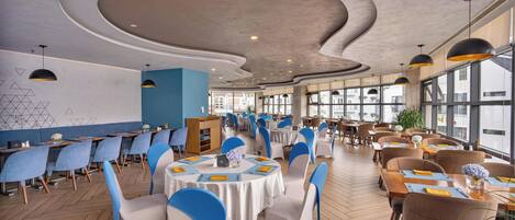 A spacious restaurant with round tables, blue and white chairs, and a modern ceiling design.