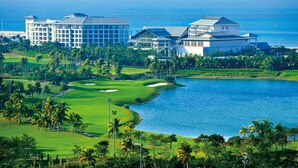 Un campo de golf con un cuerpo de agua, palmeras y un hotel con varias construcciones.
