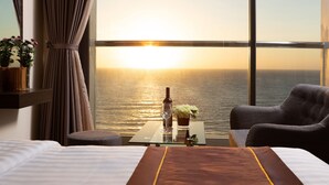 Una habitación de hotel con una cama, una mesita de vidrio con una botella y flores, una silla y vistas al atardecer sobre el agua.