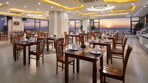 Un restaurante con mesas y sillas de madera, amplias ventanas con vistas al atardecer e iluminación de techo moderna.