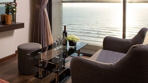 Una sala de estar moderna con una mesa de vidrio, dos sillones acolchados, una mesita de noche con un florero y vistas al mar a través de amplios ventanales.
