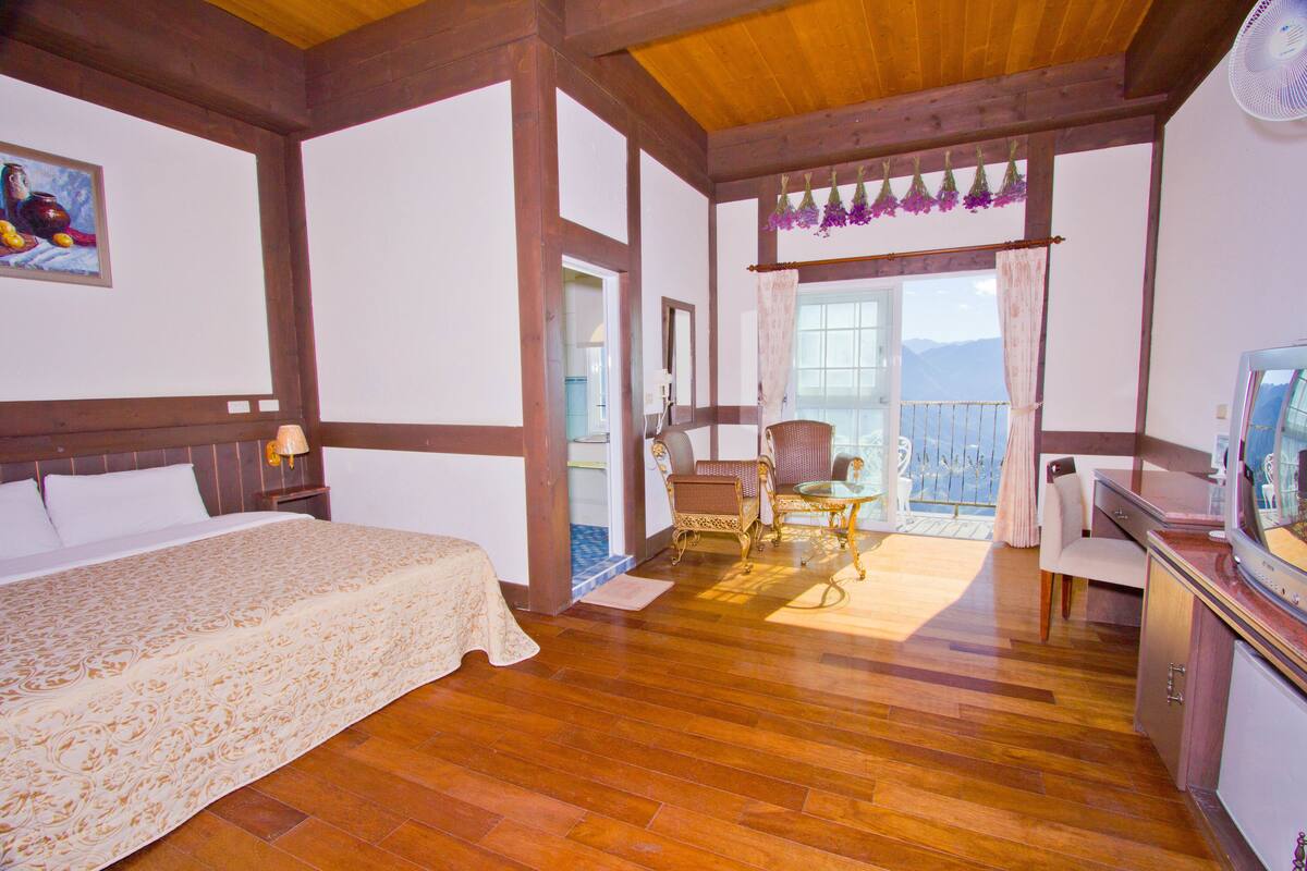 A hotel room with a wooden floor, a bed with a floral bedspread, a desk with a television, and a balcony with a view of the mountains.
