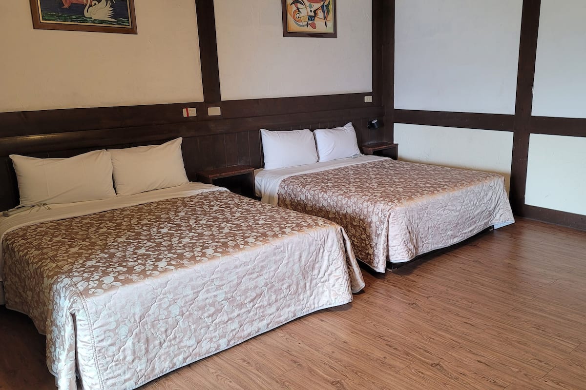 A hotel room with two beds, wooden flooring, and a small nightstand with a red item on it.
