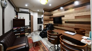 A modern living room with a brown leather sofa, a wooden dining table with chairs, a flat-screen TV mounted on the wall, and a built-in air conditioning unit. - Suzumaru Hotel - Hostel (Wakayama)