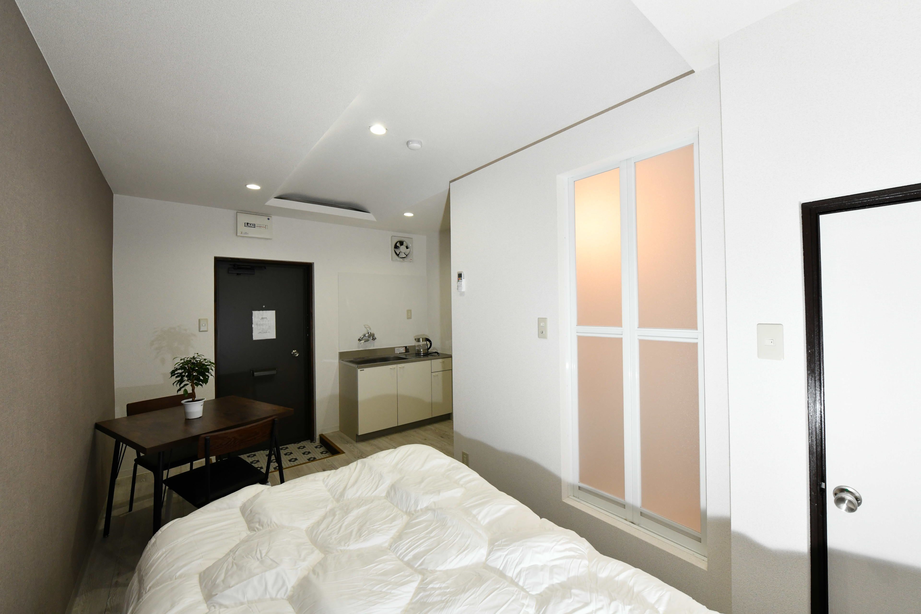 Quarto com cama, escrivaninha, cadeira, pequena mesa com planta, cozinha compacta e janela com persianas.