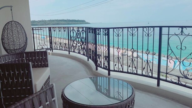 Un balcon doté d’une table et de chaises en verre, offrant une vue dégagée sur une plage, la mer et le paysage lointain.