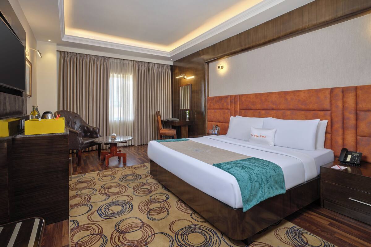 A hotel room with a large bed, a desk with a chair, a TV, and a large window with curtains.