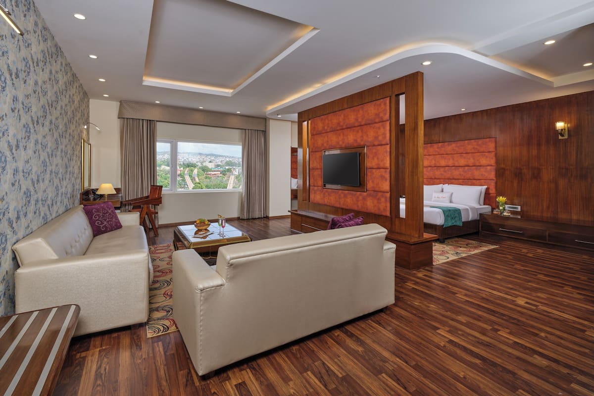 A modern hotel room with a sofa, a TV mounted on a wooden panel, a bed with a white headboard, and a view of the city through large windows.