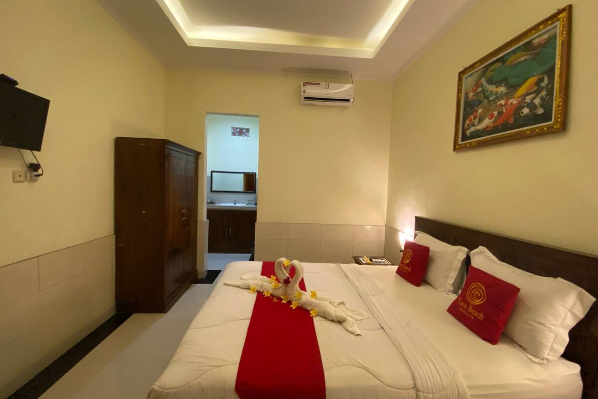 A hotel room with a neatly made bed, a red runner, a painting on the wall, and a television mounted on the wall.