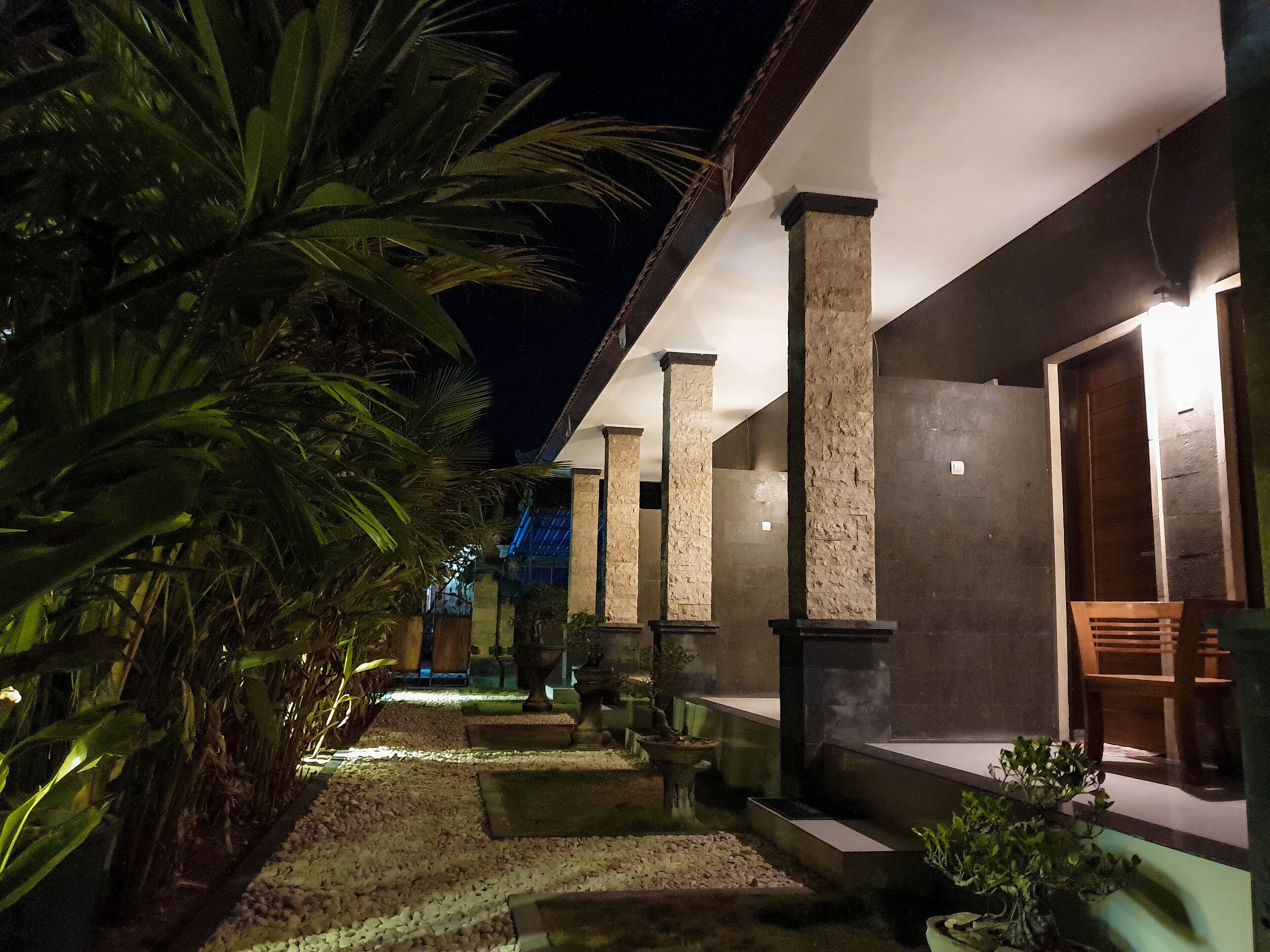 A nighttime view of a building with a pathway lined with palm trees and potted plants, leading to a covered seating area with a wooden bench and a chair.