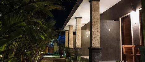 A nighttime view of a building with a pathway lined with palm trees and potted plants, leading to a covered seating area with a wooden bench and a chair.