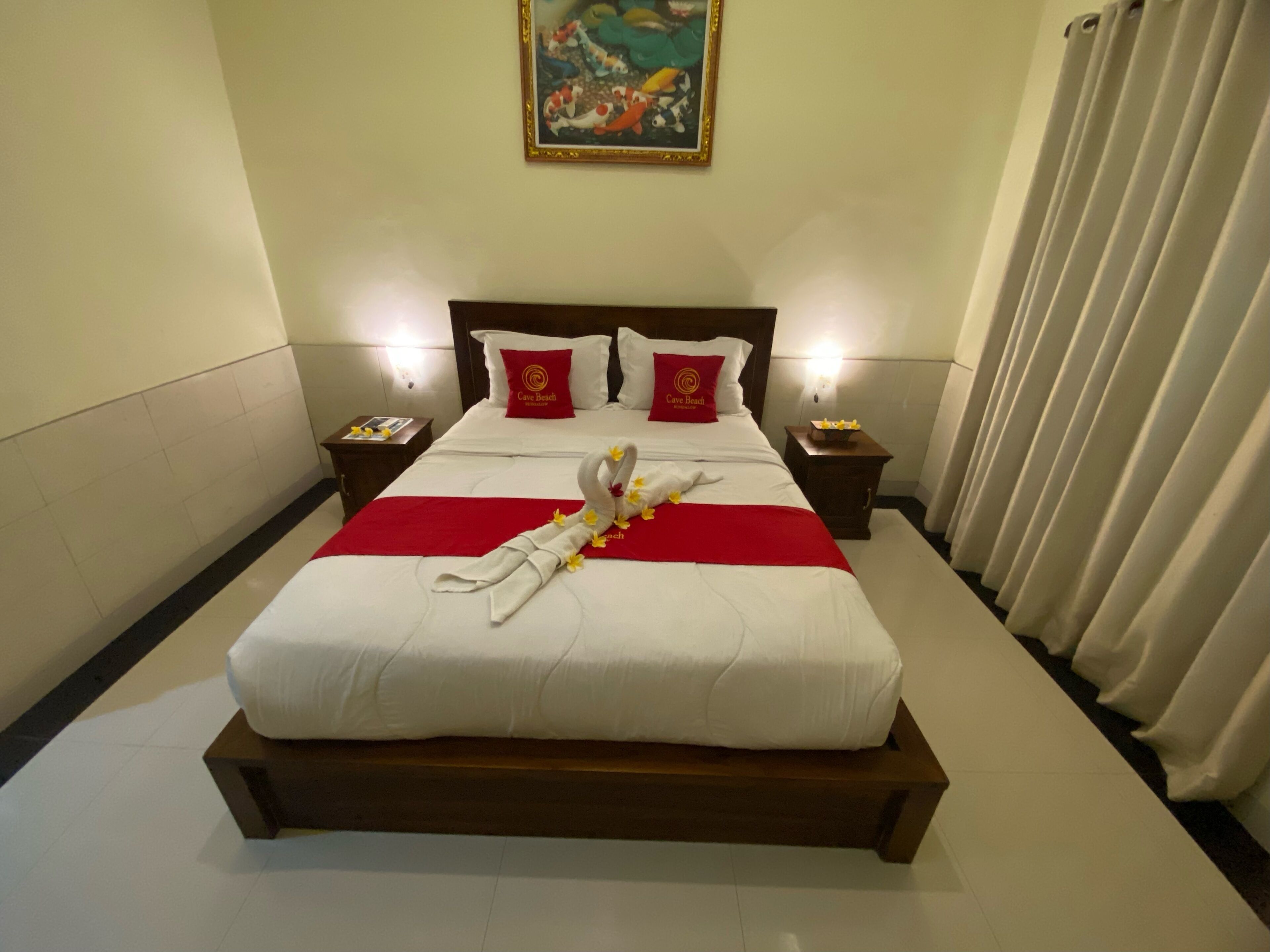 A hotel room with a large bed, two bedside tables, a nightstand, a red throw blanket, a decorative swan, and a painting on the wall.